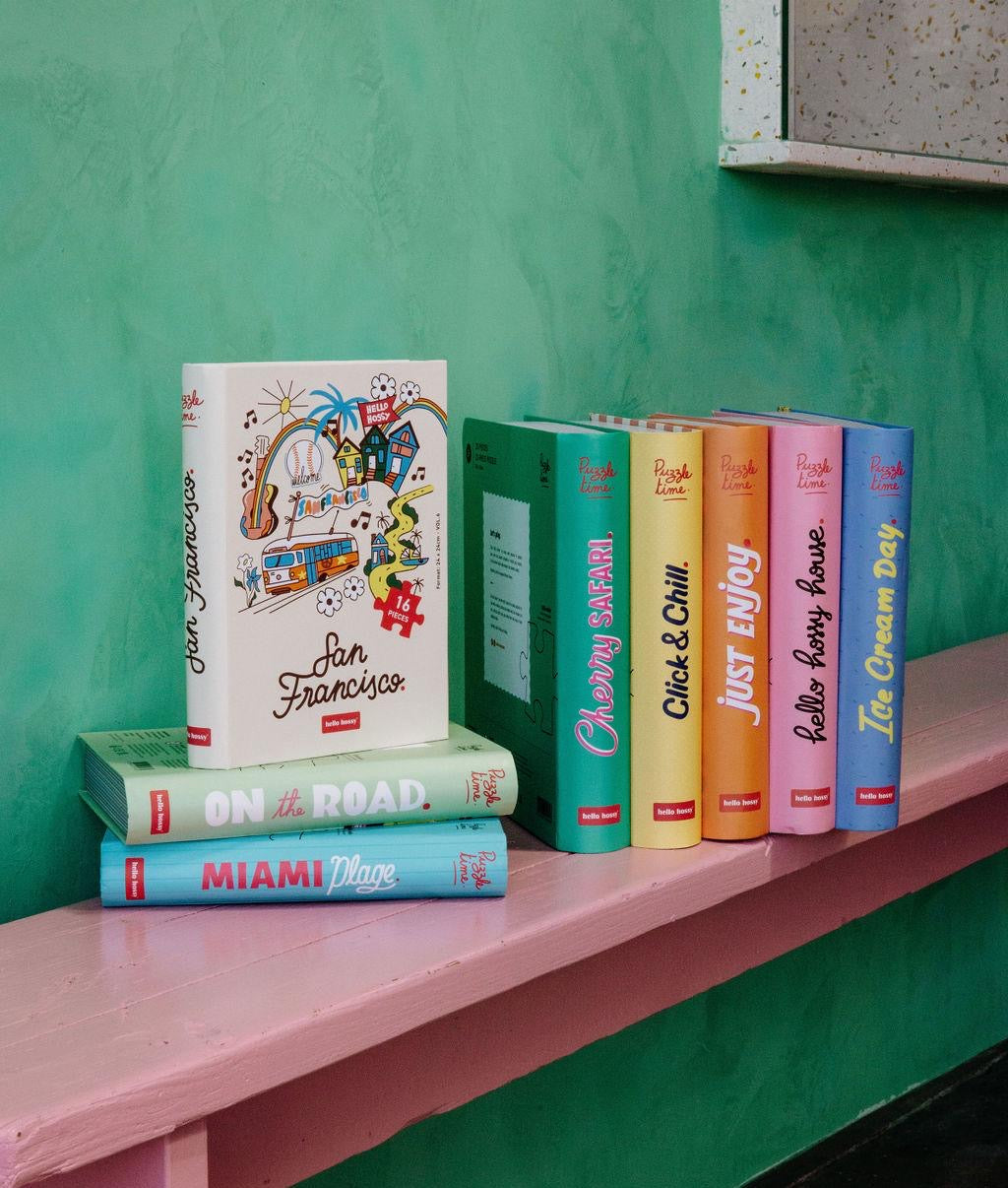 Collection de puzzles Hello Hossy posés sur un banc rose – design effet livre – fond mural coloré