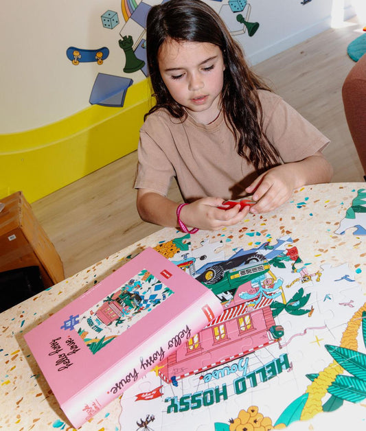 Fille qui assemble le puzzle illustré "Hello Hossy House" sur une table, avec la boîte à côté – photo lifestyle – Hello Hossy