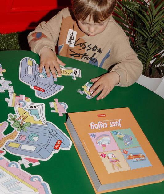 Garçon avec un des quatres puzzles "Just Enjoy"partiellement assemblé, la boîte et l'illustration modèle, sur une table, devant lui – photo lifestyle – Hello Hossy