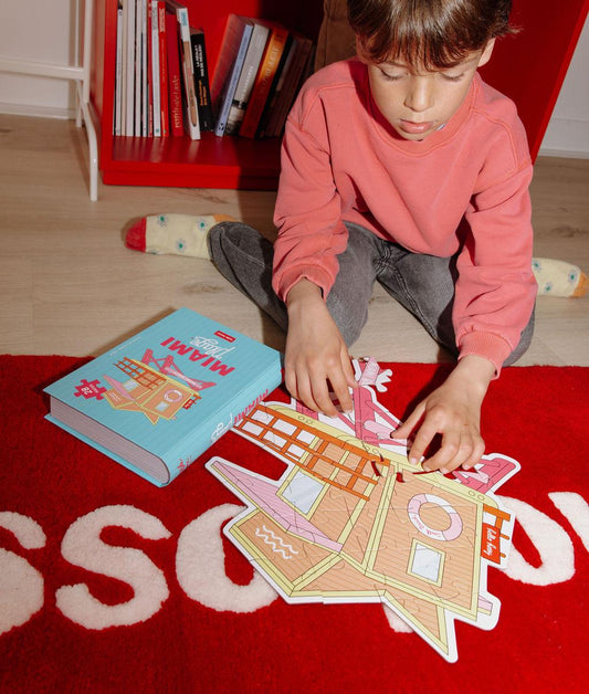 Garçon avec le puzzle "Miami Plage"partiellement assemblé et la boîte, sur un tapis, devant lui – photo lifestyle – Hello Hossy