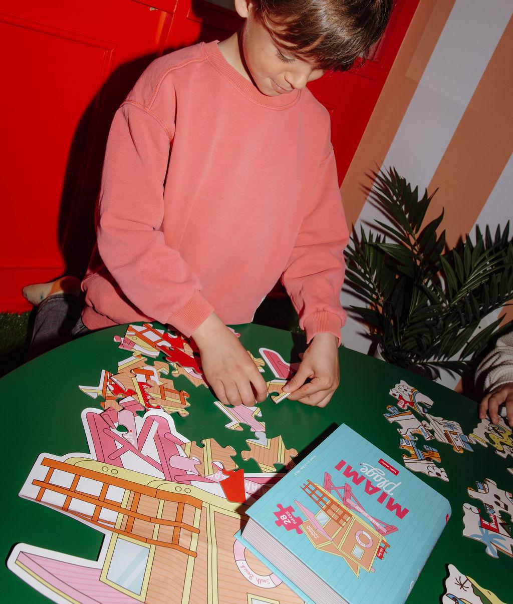 Enfant jouant avec le puzzle 28 pièces "Miami Plage" sur une table avec la boîte à côté – ambiance ludique – Hello Hossy